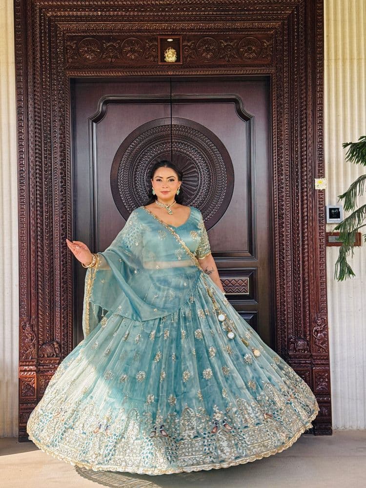Woman in a powder-blue embroidered lehenga with an organza dupatta, standing at a carved-door location.